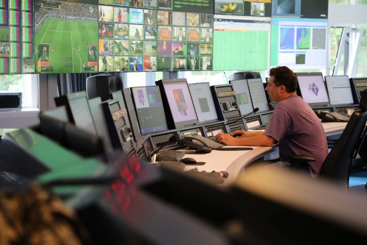 A man sitting at a curved desk backed by a long row of screens and controls. More screens are mounted behind the desk.
