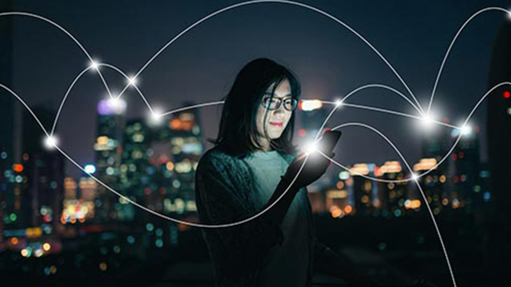 A woman using her phone with an illuminated city skyline in the distance. White lines show the digital connections being made across the city.