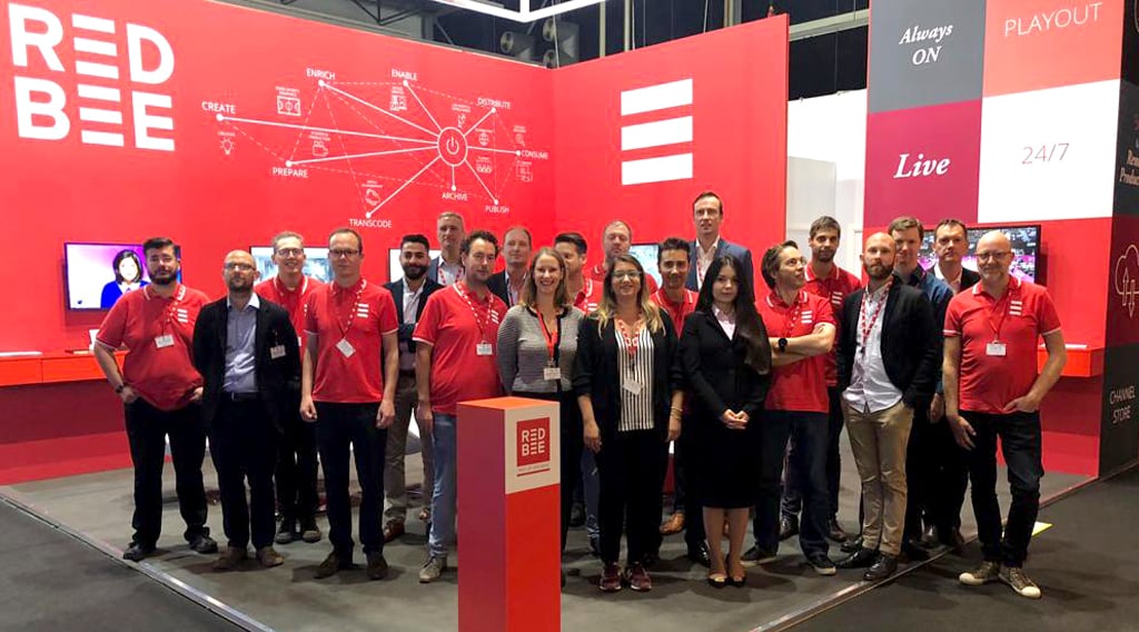 Red Bee employees posing in front of a large Red Bee stand at a convention.