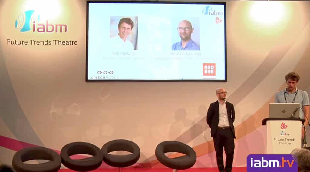 Hewson Maxwell standing under a large screen on a stage. Will Williams is standing behind a lectern next to him, looking at a laptop.