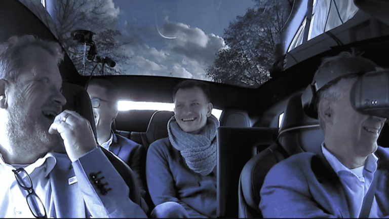 Four men sitting in a car, one wearing a VR headset.