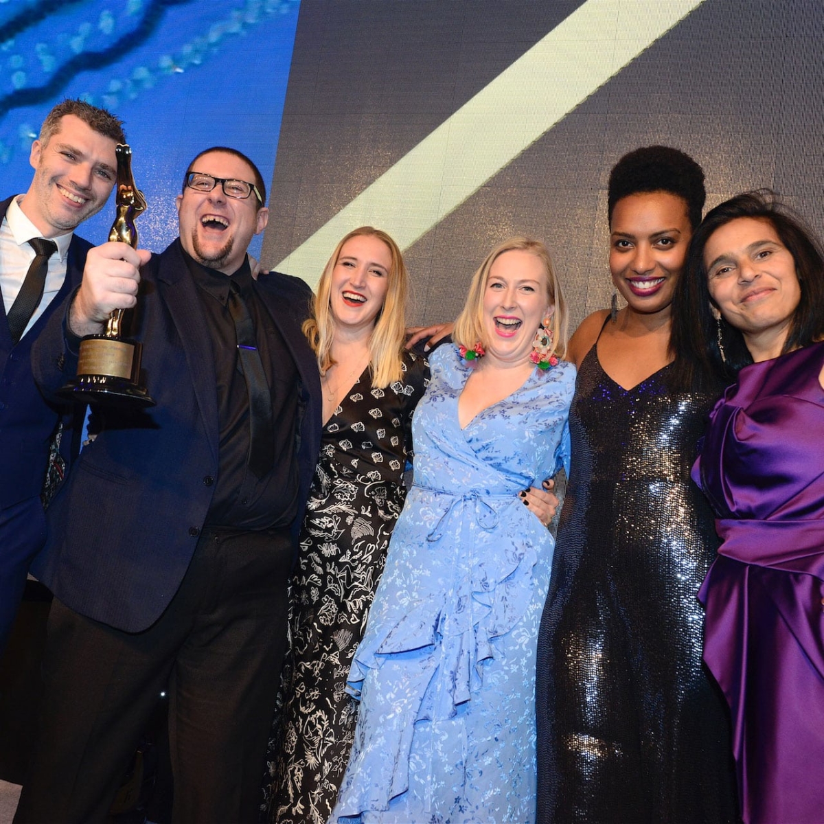 Four women and two men wearing big smiles and standing with their arms around each other at an awards ceremony. One of the men is holding up a trophy.
