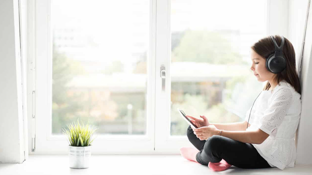 A girl sitting on a window sill, using a tablet and headphones.