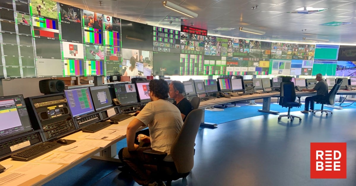 Three men sitting at a long, curved desk with screens and controls running along it. There's a wall of screens behind the desk.