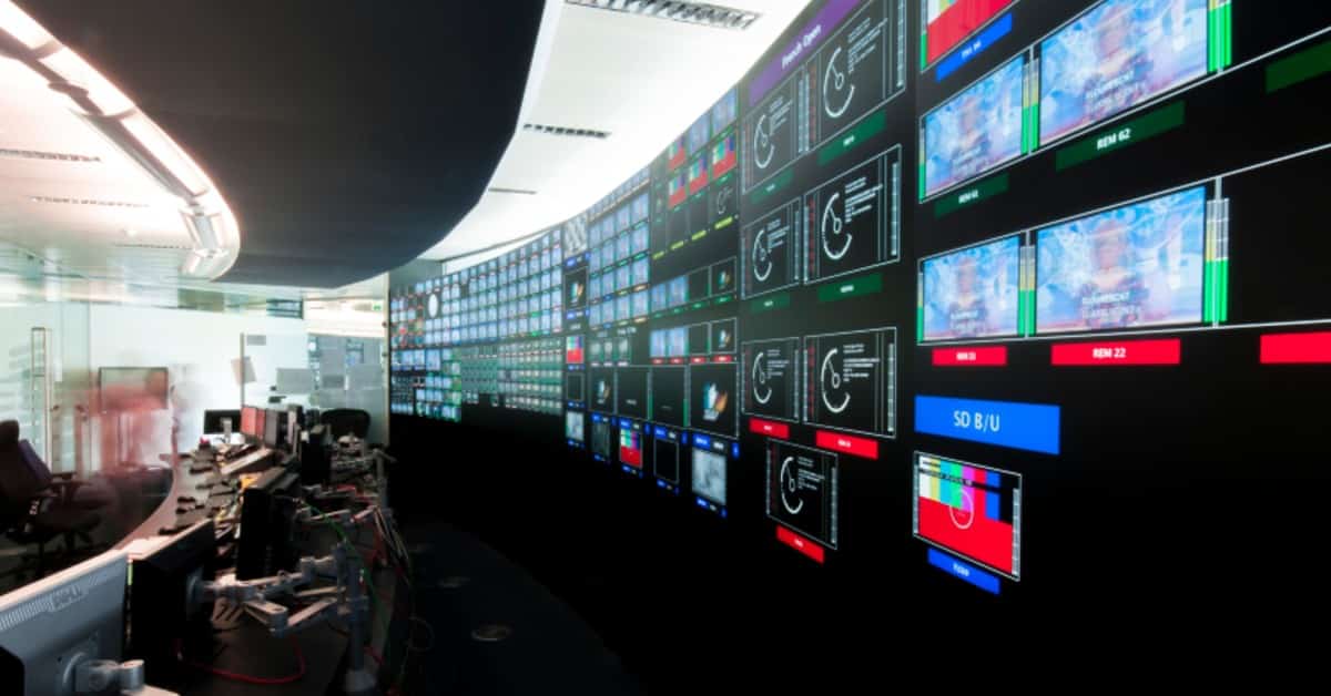A long desk with computers in front of a wall of screens.