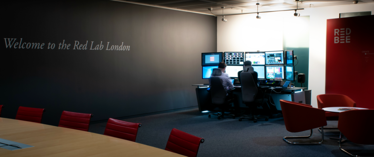 Two people sitting at a desk with two rows of screens in front of them. Welcome to the Red Lab London is painted on a wall.