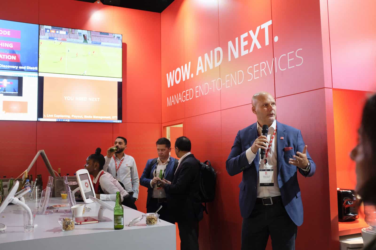 A man speaking into a microphone and standing near a table with tablet devices attached to it.