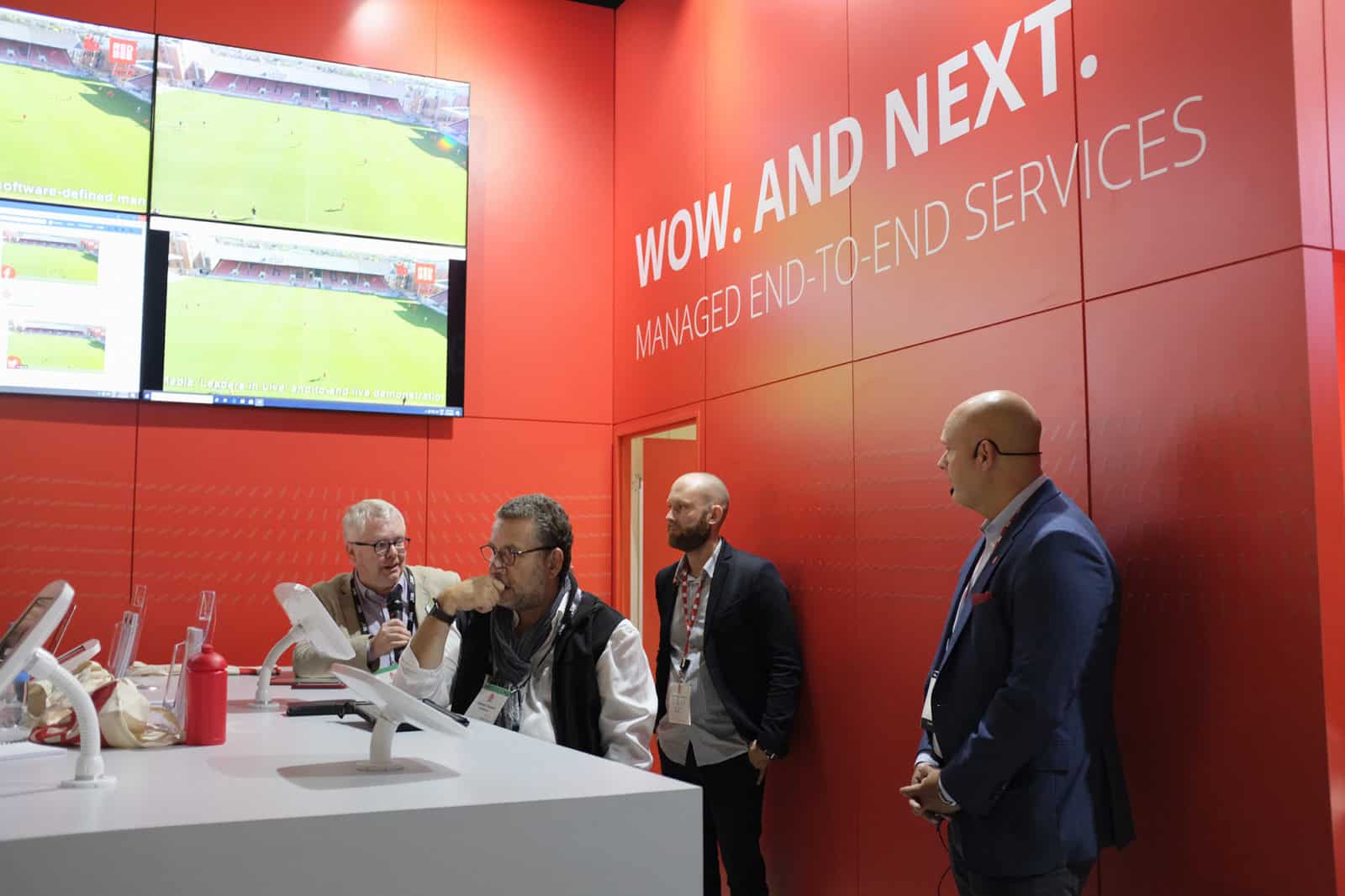Two men standing behind two other men seated at a table with tablet devices attached to it.