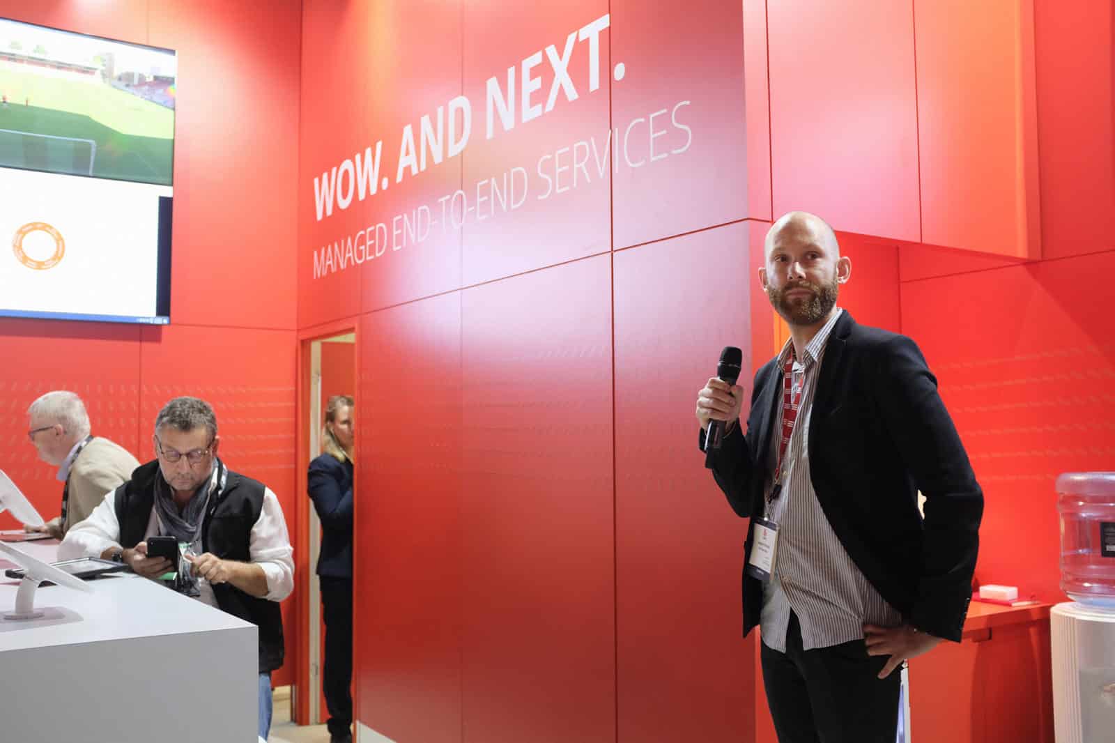 A man holding a microphone and standing near two men seated at a table with tablet devices attached to it.