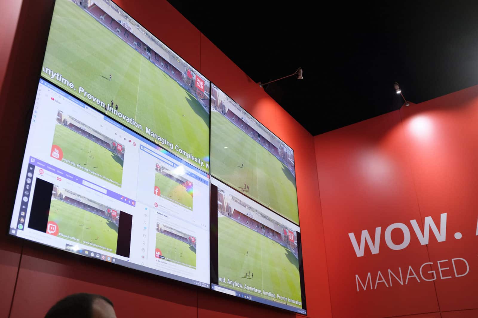 Four screens displaying a nearly empty football pitch mounted on a red wall.