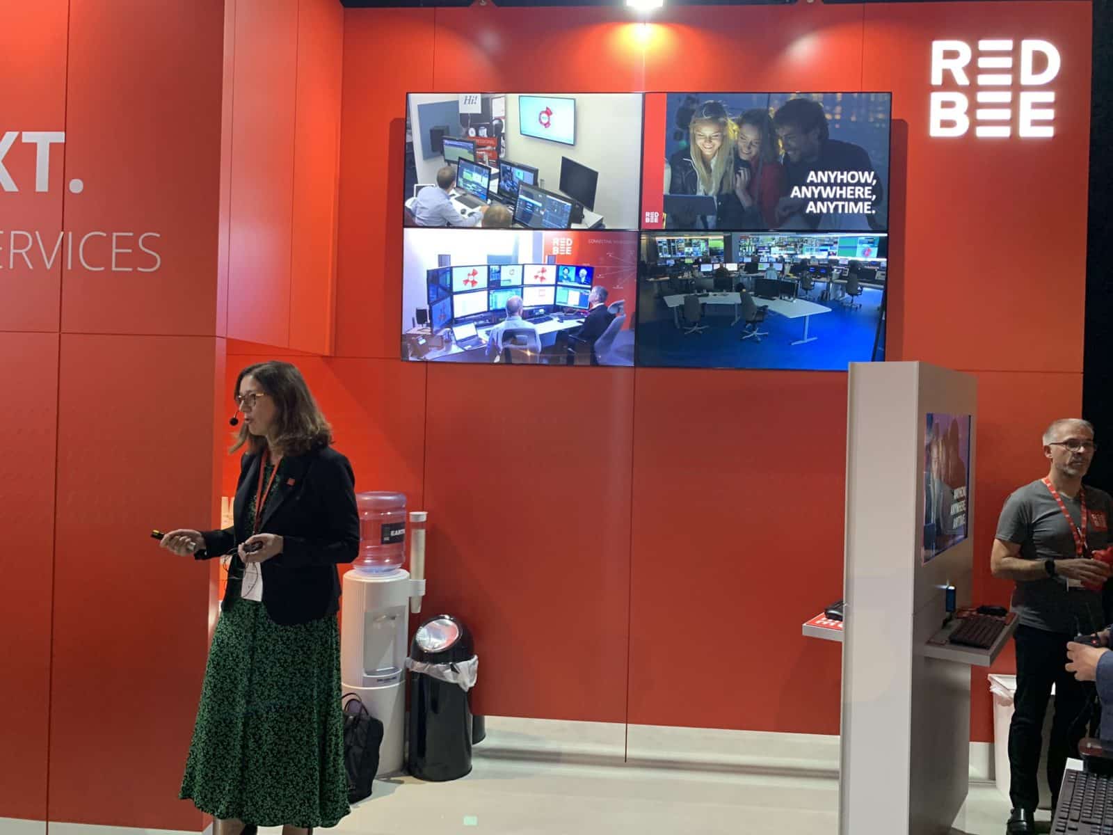 A woman wearing a headset and standing in front of a red wall with the Red Bee logo and four screens on it.