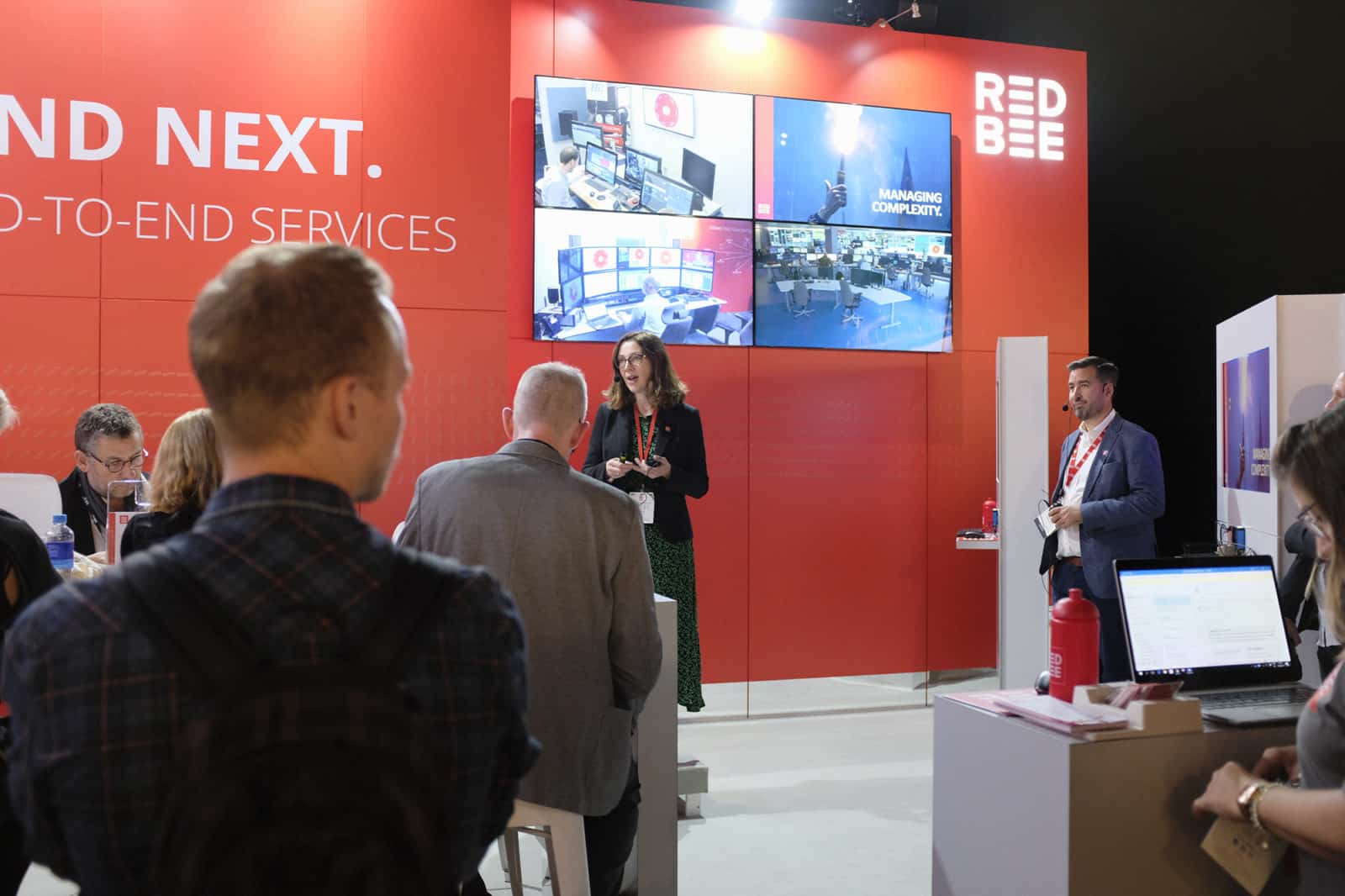 A man and a woman wearing headsets and standing in front of people seated at tables. There's a red wall behind them with the Red Bee logo and four screens on it.