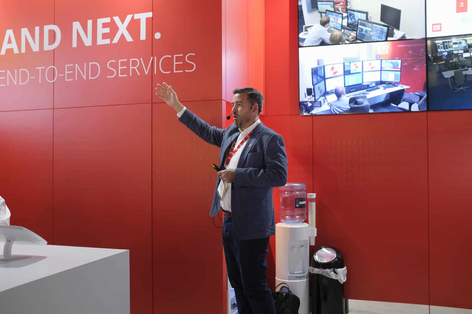 A man wearing a headset and standing in front of four screens on a red wall. He's gesturing forwards with one hand.