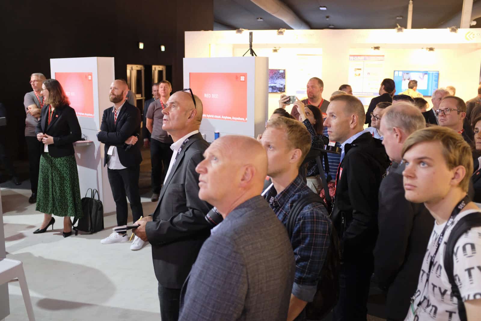 A group of people gathered near screens displaying the Red Bee logo and looking up.