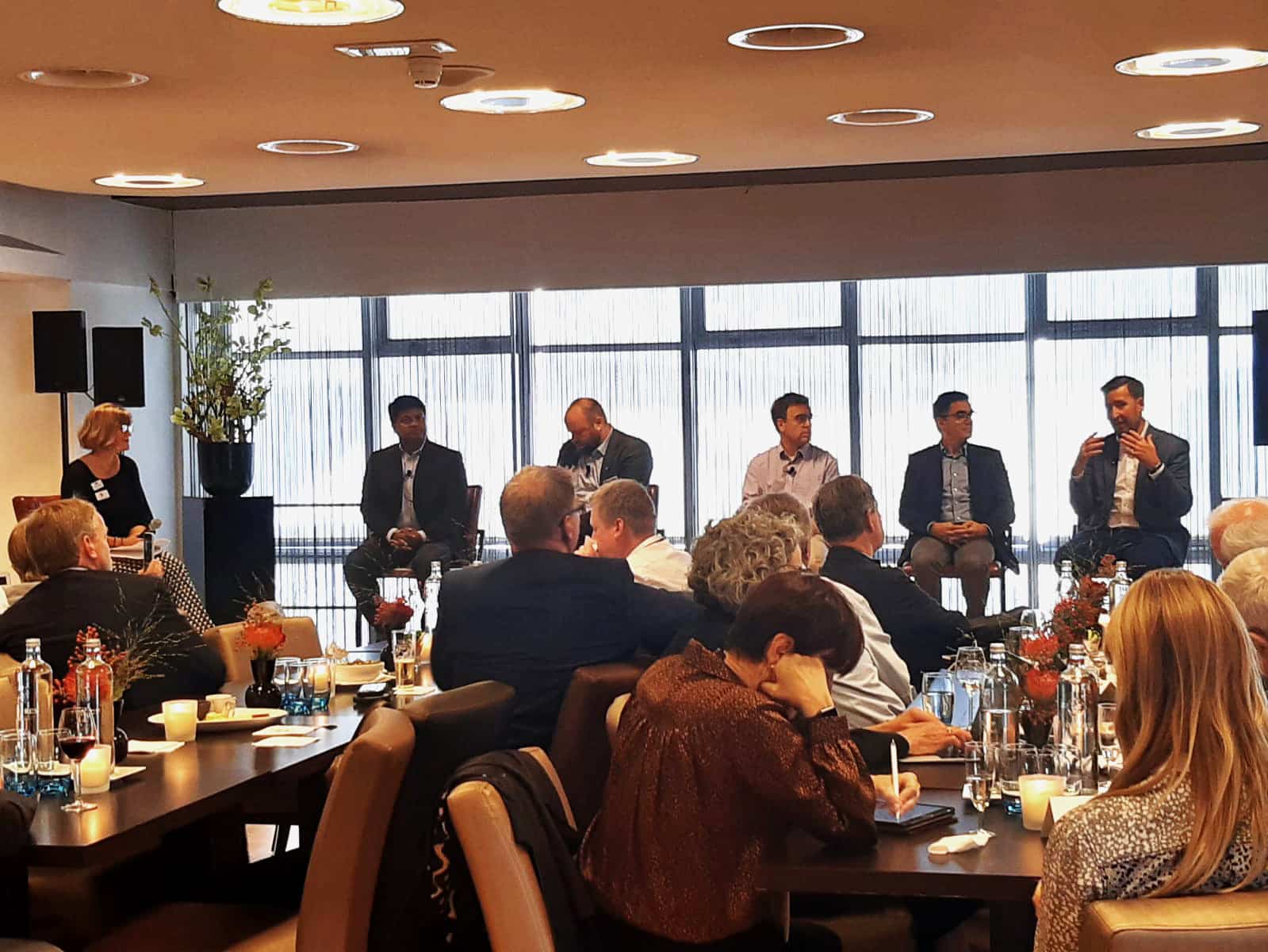 Five men sitting in a row in front of floor-to-ceiling windows. There are people sitting at long tables in front of them.