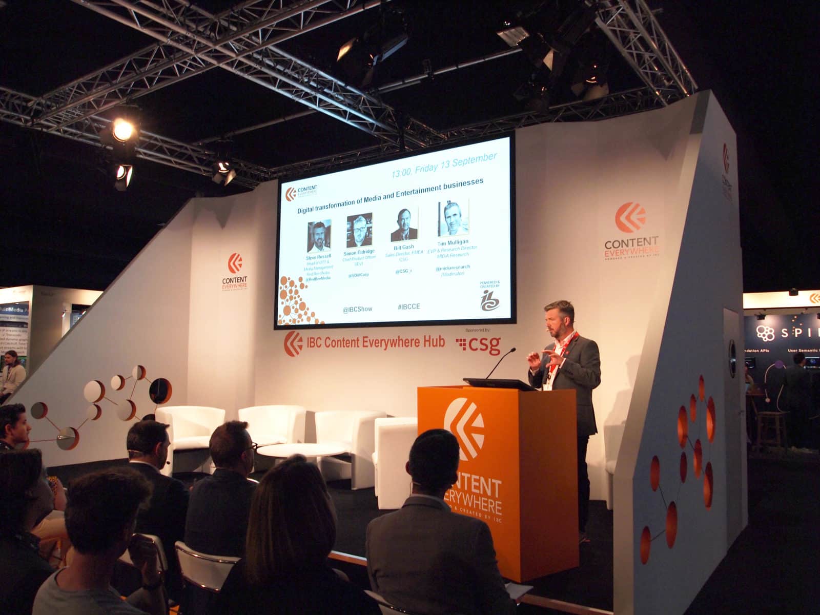 A man standing behind a lectern on a small stage, addressing people seated in rows.