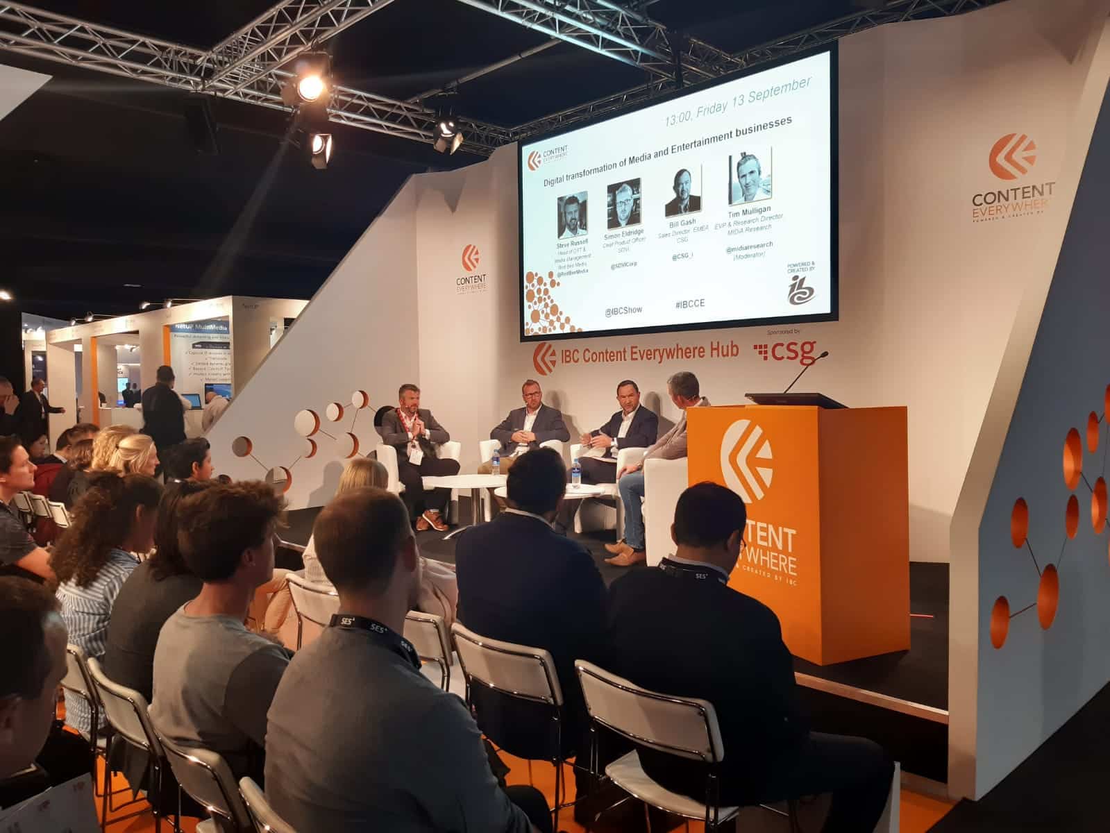 People seated in rows in front of four men sitting on a small stage.