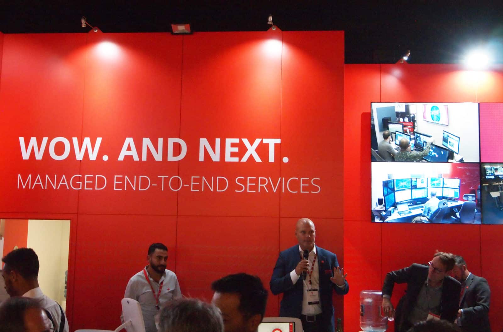 A man with a microphone standing in front of a red display wall with the white words 'Wow. And next. Managed end-to-end services' on it.