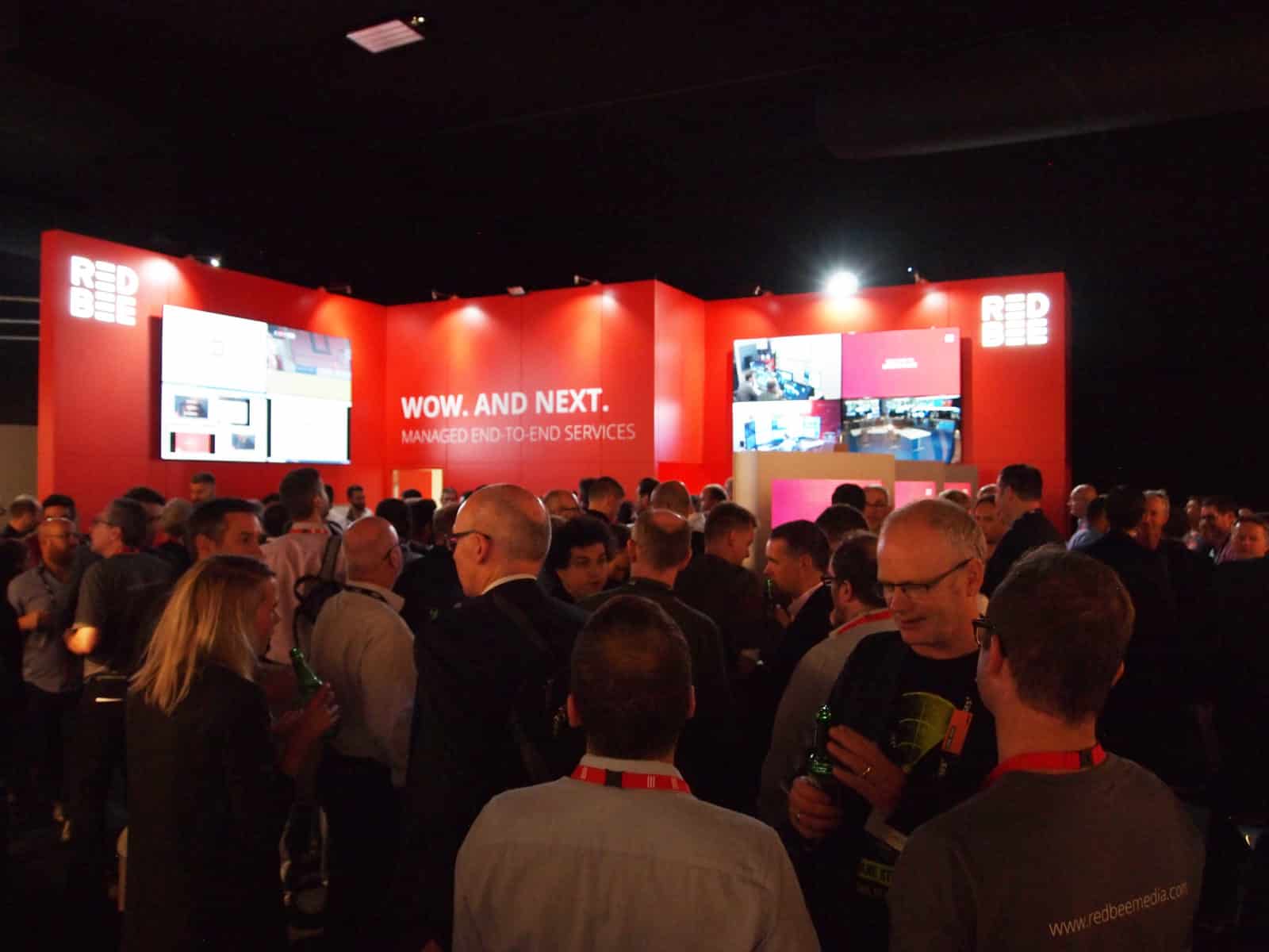 A large group of people gathered in front of a red display wall with screens and the words 'Wow. And next. Managed end-to-end services' on it.