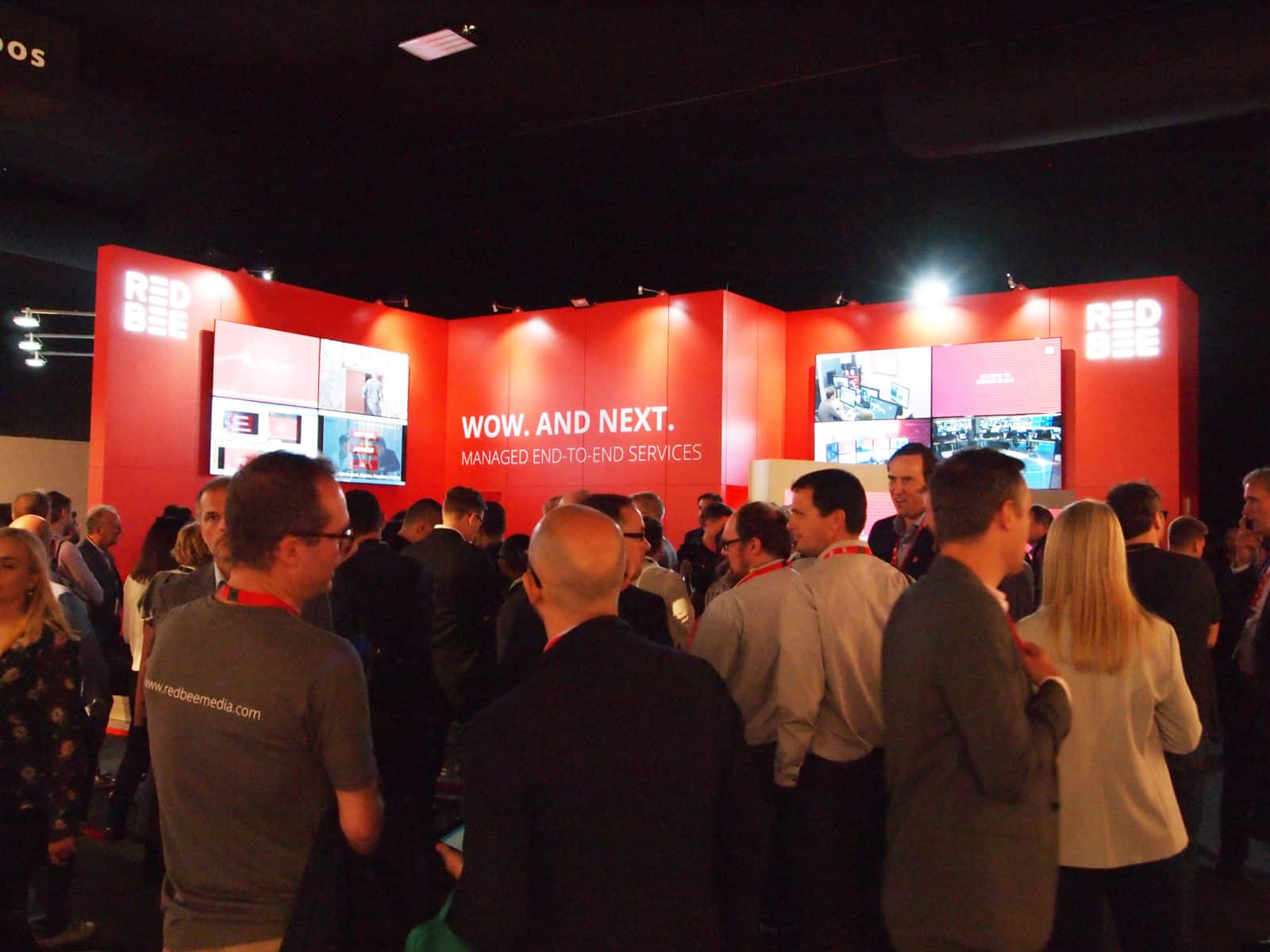 A large group of people gathered in front of a red display wall with screens and the words 'Wow. And next. Managed end-to-end services' on it.
