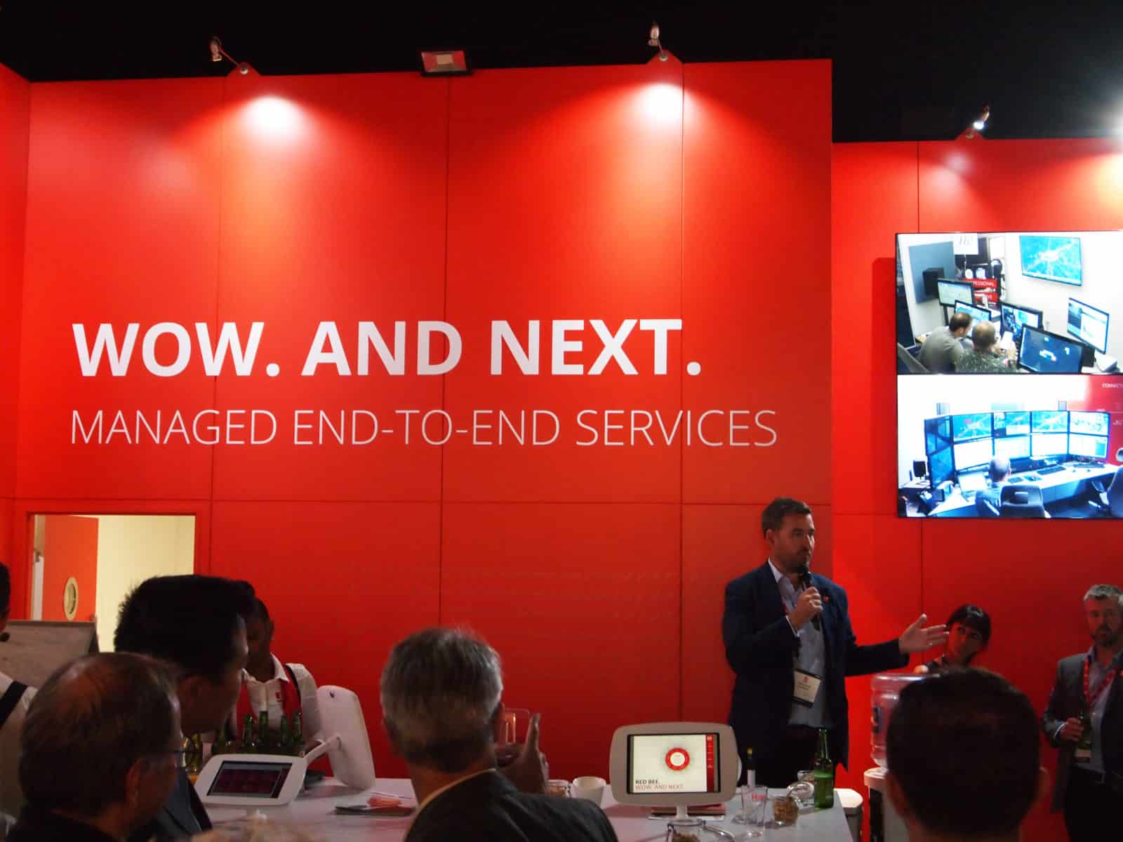 A man with a microphone standing in front of a red display wall with the white words 'Wow. And next. Managed end-to-end services'.