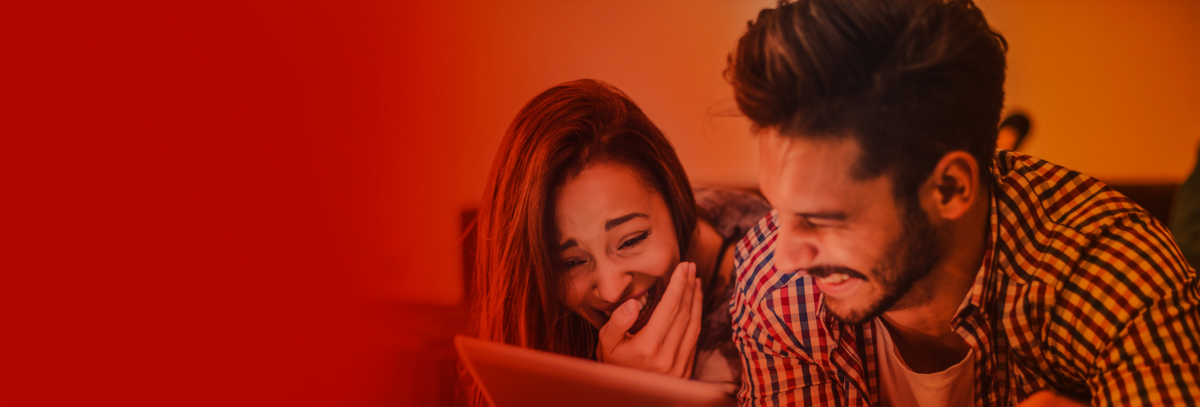 A red-and-orange tinged image showing a grinning man and woman lying on their fronts and watching something on a tablet device.