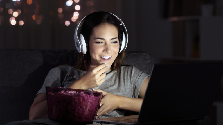 A smiling woman wearing headphones, eating popcorn, watching a laptop screen.