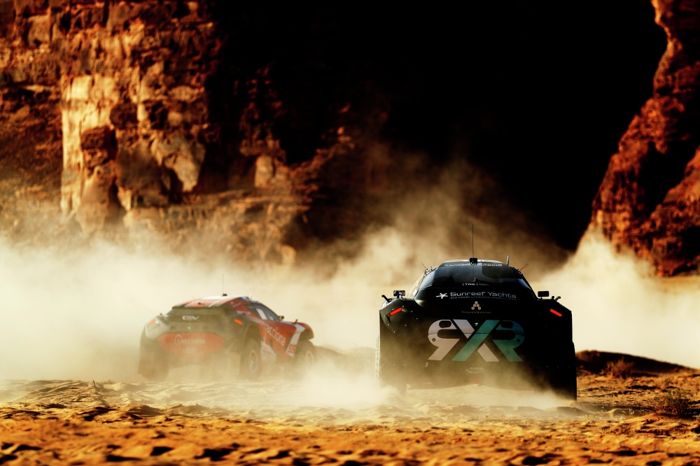 Two racing cars surrounded by a cloud of dust in a rocky, sunbaked landscape.