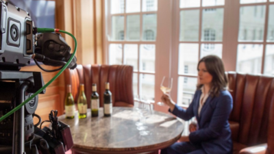 A film camera pointed at a woman sitting at a table and holding a glass of wine.