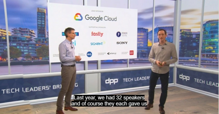 Two men standing in front of a screen displaying different companies' logos, including Red Bee's, in a television studio. A subtitle reading, 'Last year, we had 32 speakers and of course they each gave us...' is displayed at the bottom of the screen.