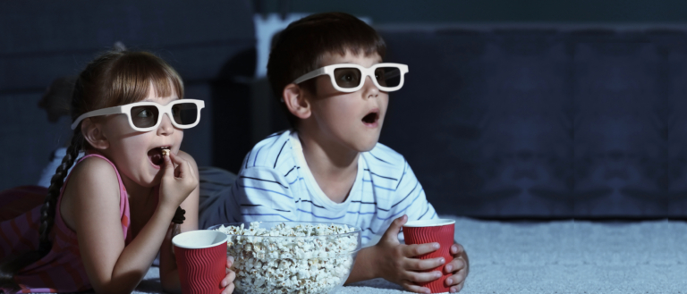 Two young children lying on their fronts and wearing 3D glasses. They're holding paper cups, and there's a bowl of popcorn between them.