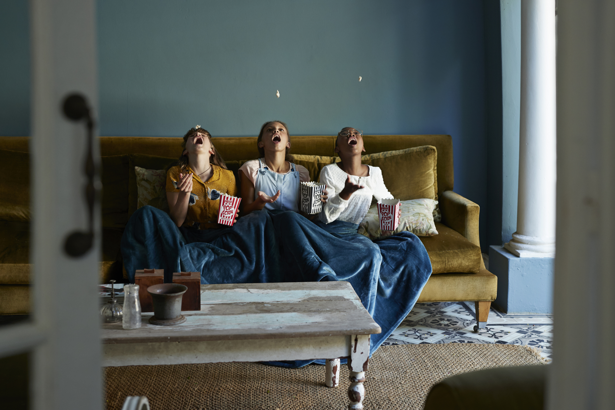 Three girls sitting on a sofa and each throwing a piece of popcorn into the air.