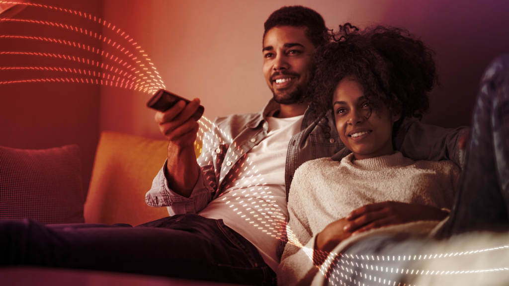 Six white-and-red, dotted lines weaving past a man holding a remote control while relaxing on a sofa with a woman.
