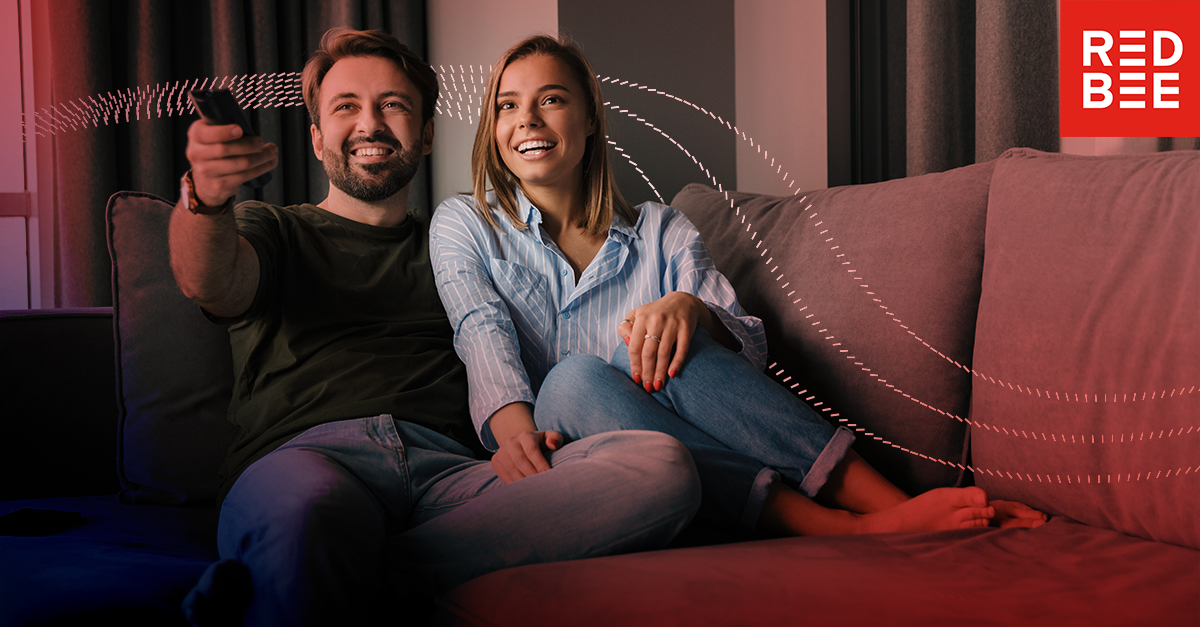 Pink-and-red, dotted lines passing behind a man and a woman relaxing on a sofa. The man's pointing a remote control.