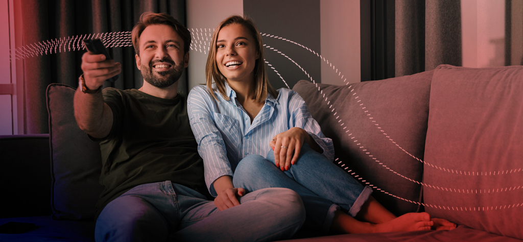 Pink-and-red, dotted lines passing behind a man and a woman relaxing on a sofa. The man's pointing a remote control.