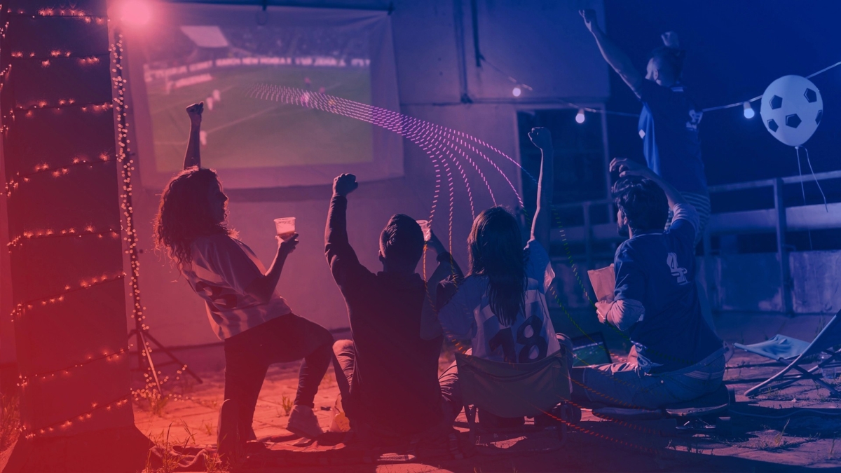 A group of football fans sit and cheer while watching a match projected on a large screen outdoors at night. They wear jerseys and hold drinks, with string lights and football-themed decorations adding to the festive atmosphere.