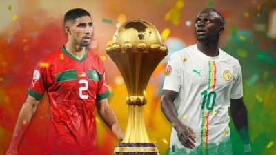 Two football players in national team kits stand on either side of a large golden trophy, with colourful confetti and a vibrant stadium-like background surrounding them.